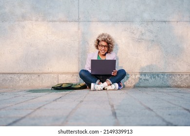 Smiling African American Business Woman Working On Laptop Sitting With Crossed Legs On Ground In City 