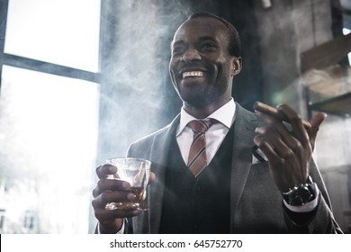 Smiling african american businessman holding glass with whiskey and smoking cigar - Powered by Shutterstock