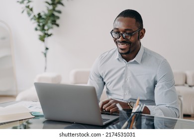 Smiling African American Businessman Wearing Glasses Working At Laptop, Makes Successful Deal. Happy Black Man Employee Glad To Receive Good News Looking At Computer Screen At Office Desk.