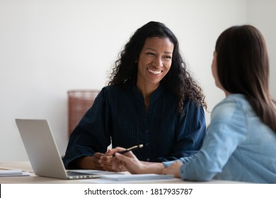 Smiling African American businesswoman talking to colleague, diverse employees brainstorming, sitting at table in office, manager consulting client, using laptop, mentor coach training intern