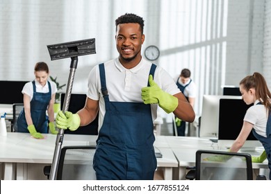 smiling african american cleaner holding vacuum cleaner brush and showing thumb up near team of colleagues
