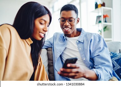 Smiling African American Couple In Love Watching Video Online On Smartphone Spending Free Time At Home, Cheerful Dark Skinned Hipster Guy Showing Gift Pictures On Mobile Phone To His Pretty Girlfriend