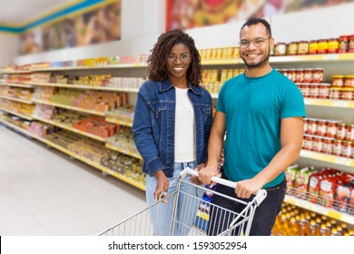 Smiling African American Couple Posing In Supermarket. Front View Of Cheerful Young People Holding One Shopping Cart While Standing In Aisle. Shopping Concept