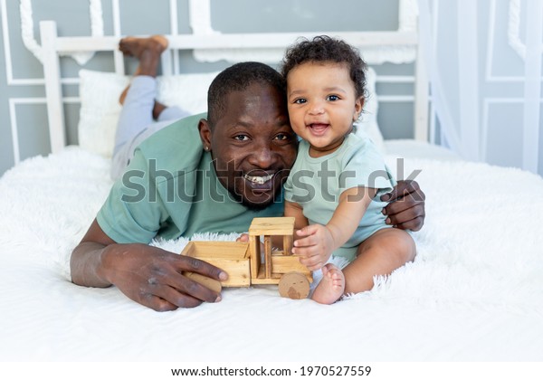 smiling\
African American dad with baby son playing on the bed at home with\
wooden toy car, happy family, father\'s\
day