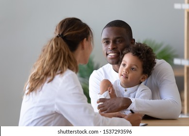 Smiling African American Dad And Little Biracial Son Visit Female Young Doctor Talking About Health Problems, Positive Ethnic Father With Small Boy Child Do Checkup Examination At Woman Pediatrician