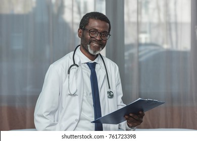 Smiling african american doctor standing with clipboard and looking at camera