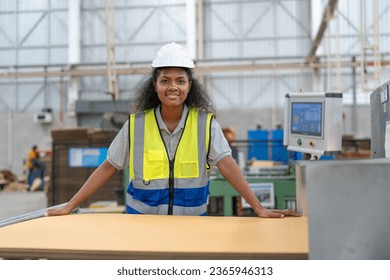 Smiling African American engineer woman in vest and helmet safety stands and works production cardboard box and packaging at factory. Female worker loading corrugated paper sheet into cutting machine. - Powered by Shutterstock