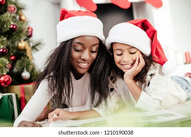 Smiling African American Family In Santa Claus Hats Looking At Photo Album In Decorated Room For Christmas At Home