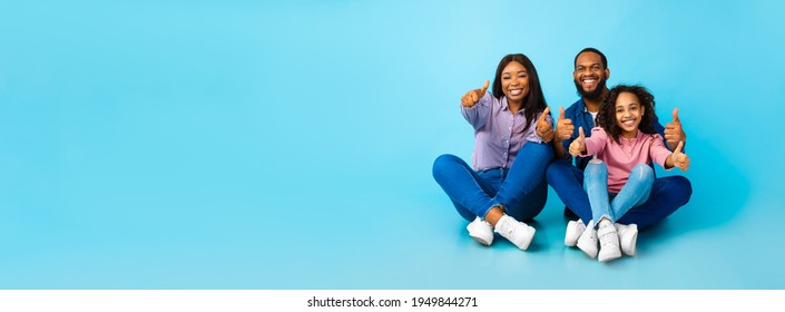 Smiling African American Family Of Three Showing Thumbs Up Gesture With Both Hands, Approving Or Recommending Something Good, Sitting On The Floor At Blue Studio Background, Panorama, Free Copy Space