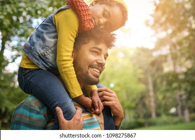 Smiling African American Father In Forest With Daughter Carrying On Shoulders.