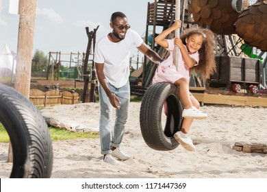 Smiling African American Father Standing Near Daughter On Tire Swing At Amusement Park