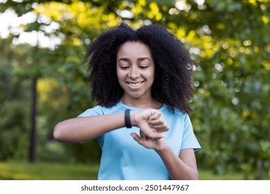 Smiling African American female athlete checks smartwatch after outdoor fitness class. She is wearing a light blue athletic shirt and stands in a lush green park . Health and fitness concept. - Powered by Shutterstock