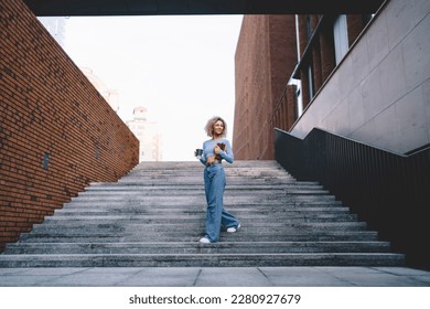 Smiling African American female in casual clothes walking on stone stairway with paper coffee cup and diary in hands and looking away near brick wall on city street