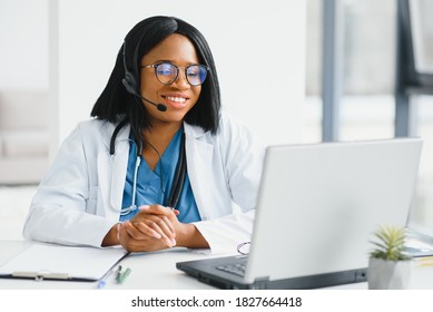 Smiling African American Female Doctor Gp Wears White Medical Coat Using Laptop Computer At Workplace Gives Remote Online Consultation, Working On Pc, Consulting Patient In Internet Telemedicine Chat.