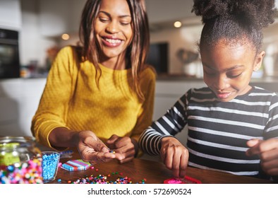 Smiling African American Female Parent In Yellow Sweater Working With Cute Child In Striped Shirt On Beading Project At Table In Kitchen