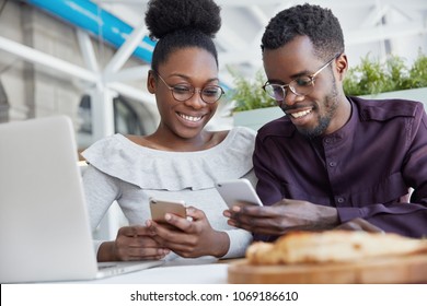 Smiling African American friends meet together at cafe, use modern technologies for entertainment. Dark skinned delighted young female and male hold smart phones, download new applications or files - Powered by Shutterstock