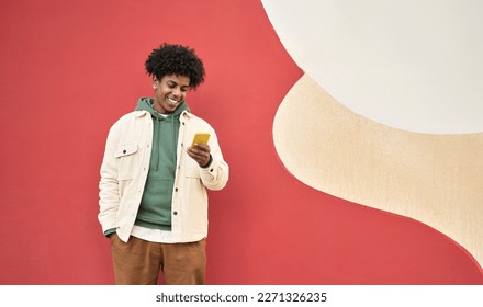 Smiling African American gen z teen standing at color wall using mobile phone. Happy cool ethnic stylish hipster guy model holding cell phone, checking apps on mobile device, looking at cellphone. - Powered by Shutterstock