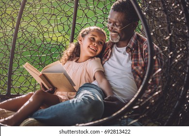 smiling african american girl reading book while sitting in swinging hanging chair with granddad