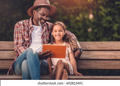 Smiling African American Grandchild And Her Grandfather Listening Music On Digital Tablet While Sitting On Bench In Park  