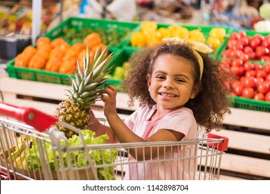 smiling african american kid sitting in shopping trolley with pineapple in grocery store 
