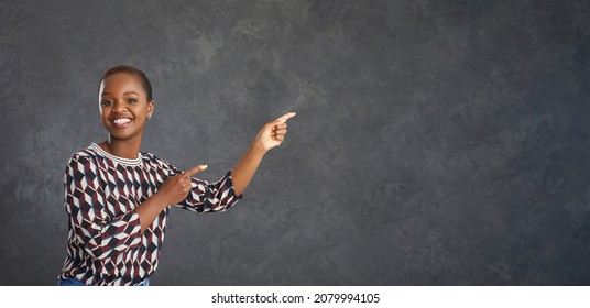 Smiling African American lady with short haircut promoting new product. Happy attractive young black woman in modern geometric pattern jumper points index fingers away at grey copy space background