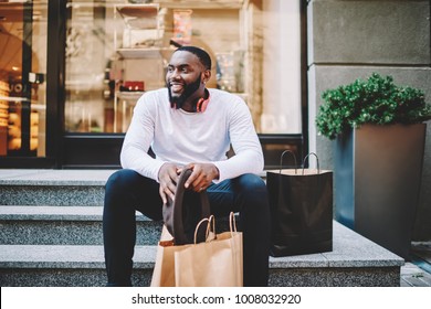 Smiling African American Male Customer In Trendy Wear Sitting On Stairs Of Store With Bags With Copy Space For Label, Cheerful Dark Skinned Hipster Guy Recreating After Shopping And Buying Purchases