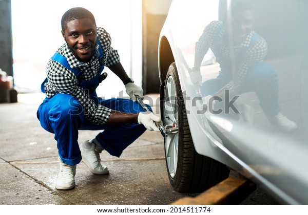Smiling african\
american male mechanic in a car service sitting near a car wheel\
and changing a tire using a\
wrench