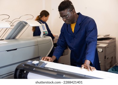 Smiling African American male specialist in a blue robe uniform pressing the buttons on the plotter in the typography - Shutterstock ID 2288421401