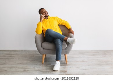 Smiling African American Male Talking On Cellphone Sitting In Armchair Near Gray Wall At Home. Black Guy Having Phone Conversation Via Smartphone, Chatting Relaxing In Chair Indoor. Communication