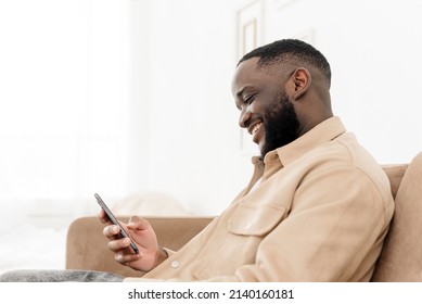 Smiling African American Man Browsing News Or Social Media Feed On Phone While Sitting On Sofa At Home. Rest And Relaxation