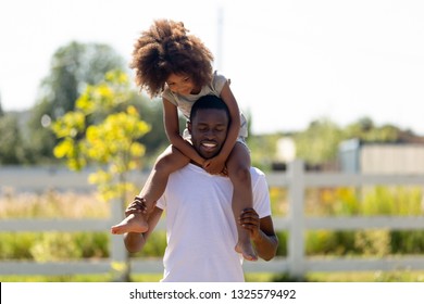 Smiling African American Man Carrying Preschool Daughter On Shoulders, Playing Funny Game Outdoor Together, Enjoying Weekend, Family Activity, Happy Man Walking With Little Girl In Yard