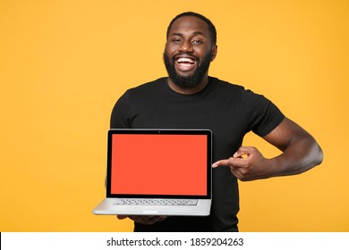 Smiling African American Man In Casual Black T-shirt Isolated On Yellow Background. People Lifestyle Concept. Mock Up Copy Space. Pointing Index Finger On Laptop Pc Computer With Blank Empty Screen