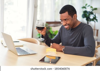 Smiling African American Man Celebrating Birthday At Home During Pandemic Quarantine Time. He Drinking Red Wine And Cake With Friends And Family On Through Video Call Virtual Party With With Laptop.