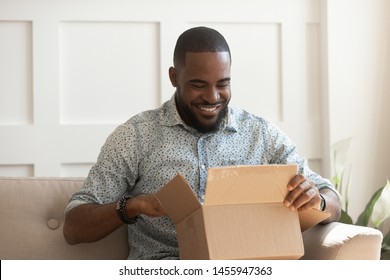 Smiling African American Man Consumer Open Cardboard Box Get Postal Parcel, Happy Black Male Customer Receive Carton Package Sit On Sofa At Home Satisfied With Fast Shipment Online Purchase Delivery