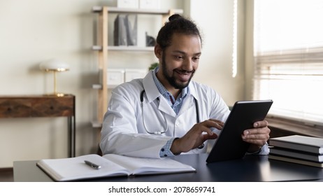 Smiling African American Man Doctor In White Uniform With Stethoscope Using Tablet, Sitting At Desk In Office, Physician General Practitioner Looking At Device Screen, Consulting Patient Online