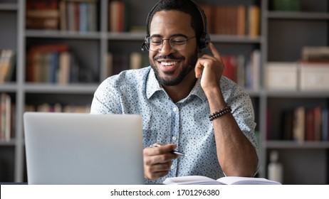Smiling African American man in glasses and headset watch webinar on laptop making notes, happy biracial male student worker in headphones handwriting studying or working using computer