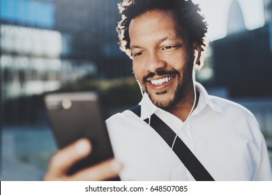 Smiling African American Man In Headphone Making Video Call Via Mobile Phone In Hand.Concept Of Guy Using Internet-enabled Electronic Device Outdoor.Blurred Background