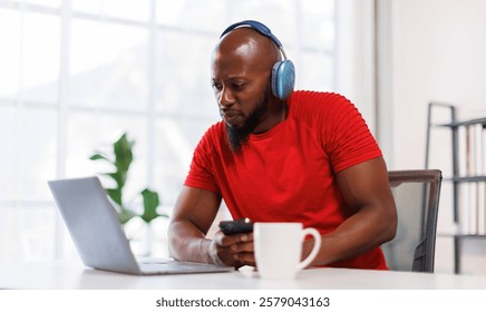 Smiling african american man in headphones laptop and typing something on phone at home workplace.