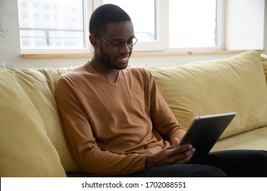 Smiling African American Man Holding And Using Tablet With Social Networks At Home, Happy Black Freelancer Making Web Shopping, Sitting On Cozy Sofa At Living Room.