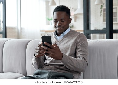 Smiling African American Man Holding Smart Phone Using Social Media Apps Sitting On Couch At Home. Black Guy Texting, Remote Learning, Ordering Distance Delivery Or Communicating Online In Application