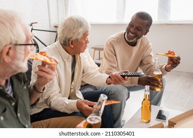 Smiling african american man holding beer and pizza near multiethnic friends with remote controller at home
