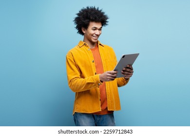 Smiling African American man holding digital tablet ordering food, shopping online isolated on blue background. Happy modern hipster using mobile app, watching videos, rearing e-book. Technology - Powered by Shutterstock