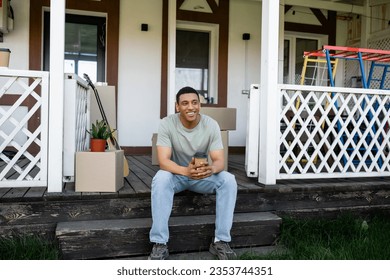 smiling african american man holding takeaway coffee while sitting on porch of new house - Powered by Shutterstock