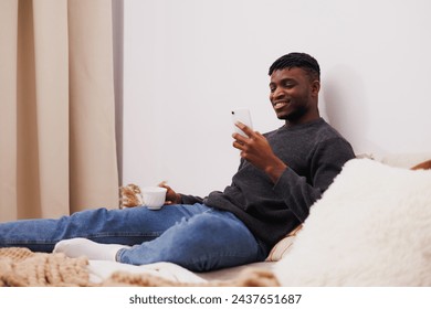 Smiling african american man in jumper using smartphone and holding coffee on bed - Powered by Shutterstock