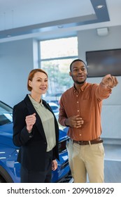 Smiling African American Man Looking Away And Pointing With Finger Near Car Dealer