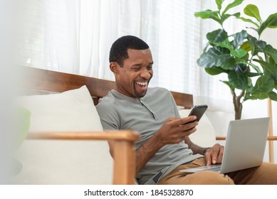 Smiling African American man making online video call on mobile phone while working on laptop. Black male enjoy with internet technology while talking and chatting with family on notebook at home