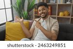 A smiling african american man points at an imaginary object in his open hand, sitting in a cozy home interior.