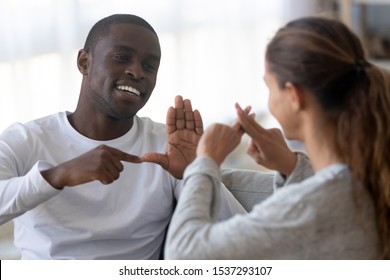 Smiling african American man sit on couch show hand gestures talking with female friend at home, international disabled hearing impaired couple or spouses use sign language communicating