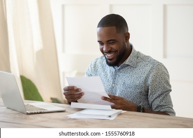 Smiling african American man sit at desk working at laptop reading good news in postal correspondence letter, happy biracial male feel excited get pleasant positive response reply in post notice
