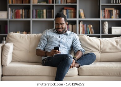 Smiling African American Man Sit Relax On Comfortable Couch At Home Using Modern Cellphone, Happy Biracial Millennial Male Rest On Sofa In Living Room Texting Messaging On Smartphone Gadget